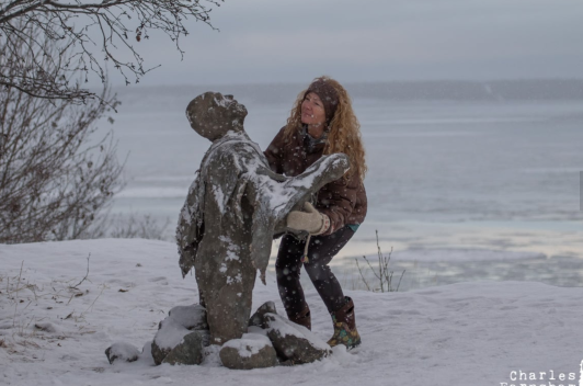 Person leaning down to hug a stone statue of a person in the snow by a lake.