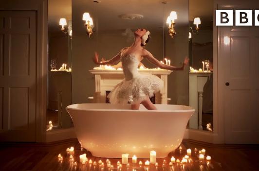 In a bathtub surrounded by candles, stands a ballet dancer in a white tutu posing.