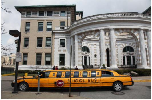 A limo sits in front of a white and beige building. The limo is painted yellow and black with the words "school bus" on it.
