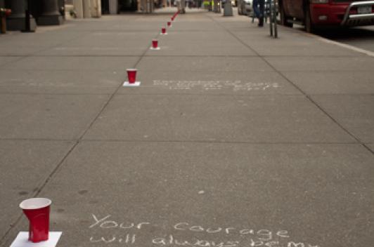 On an empty sidewalk in the city, red solo cups sit about 6 feet apart with words written beside them in white chalk.