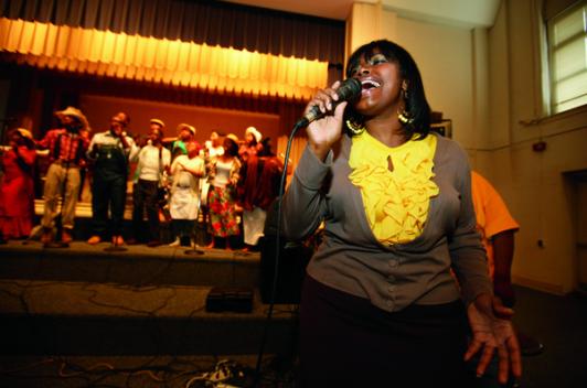 A person standing and singing with a group behind them accompanying them