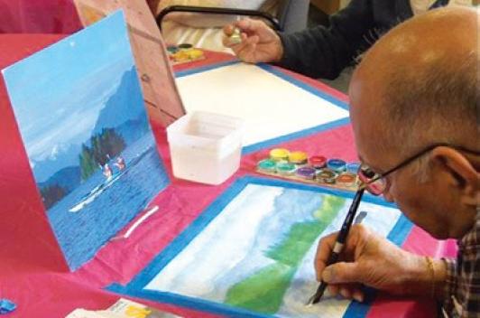 An older person sits at a table with a red table cloth. They are painting a landscape.