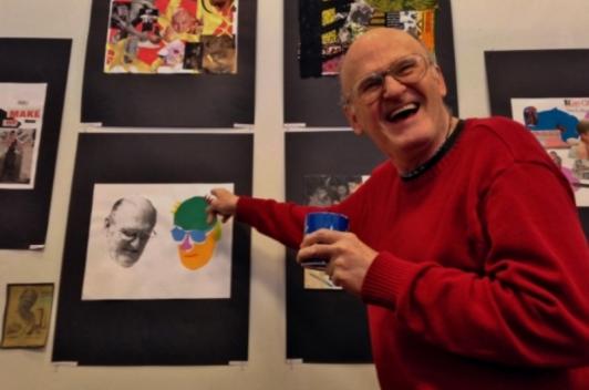 A person in a red sweater smiling and point at a work of art featuring his own face