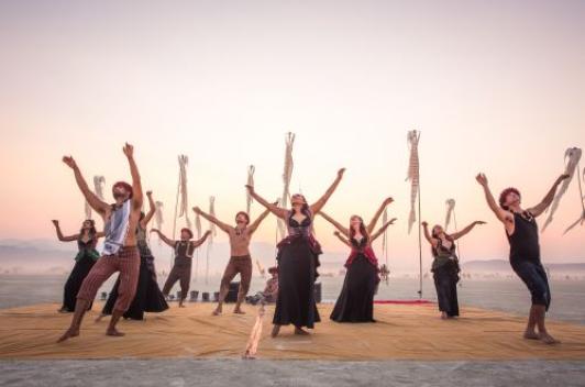 A group of dancers standing in the desert with the sun behind them. They are all in black and are swing pieces of fabrics.