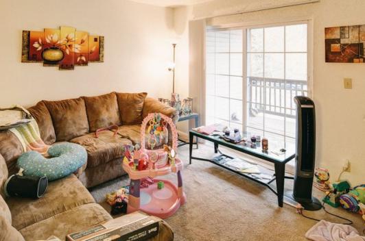 A living room with two tan couches and various wall art. There is a sunny sliding door that leads to a balcony. A baby bouncer sits in the middle of the floor.
