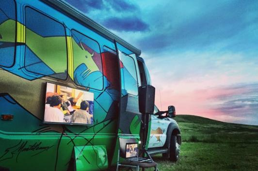 A bus has a painting of abstract animals with a tv attached to the side. It is sitting on a lawn during sunset.