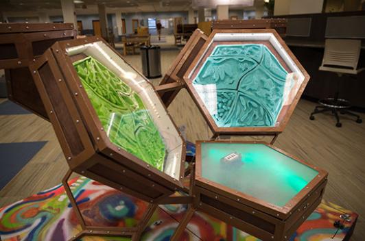 A sculpture that features hexagons made of metal with glass fronts. Inside are green and blue sculptures with ridges, hills and valleys.