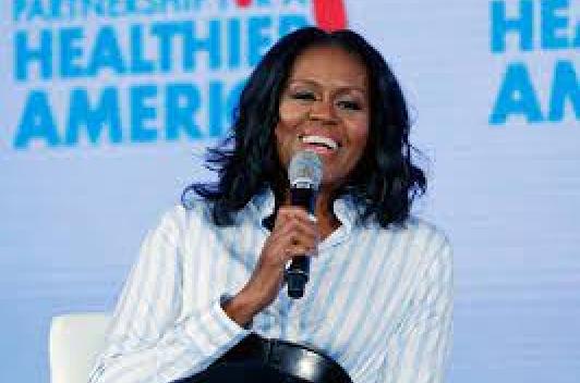 A smiling Michelle Obama sits in a white chair in front of a blue screen, speaking into a mic.