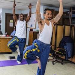 Two people in white tank tops and blue pants doing yoga. A prison guard sits behind them at a desk