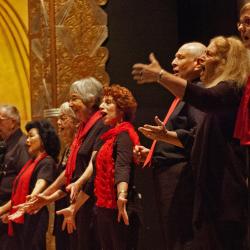 A group of older people standing on a stage in black outfits with red scarfs, singing