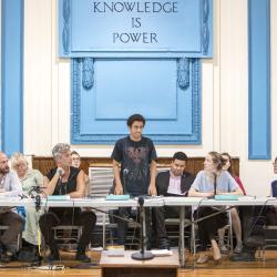 People of various ages sitting at a table in front of a wall with the words Knowledge is Power. One person is standing with a determined look on their face