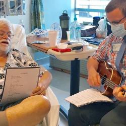 Person with a ukulele sitting with another person in a hospital bed who is singing along