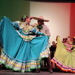 Couples dancing in front of a projection of the Mexican flag. Some a wearing large skirts that they are holding wide, while others are in hats