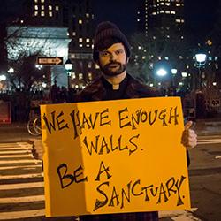 A person standing near a crosswalk holding a sign that says, "We have enough walls. Be a sanctuary."