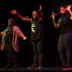 Three people standing on a stage in front of mics performing spoken word poetry