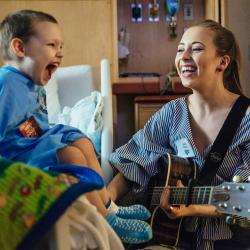 Person with a guitar singing along with a child sitting in a hospital bed. Both are in blue.