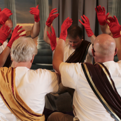 People sitting in a circle in robes with sashes holding up their hands. Their hands have red gloves on them