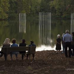 People standing and sitting by a lake that has thin poles emanating light coming up from the water.