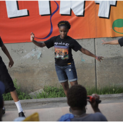 Three dancers stand on a platform, dancing in sync. Behind them is a colorful banner with the word, Flint, on it.