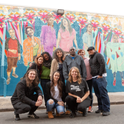 A small group gathered in front of a mural posing and smiling.