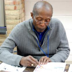 An older person in a grey sweater sits at a table drawing.