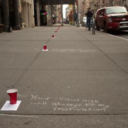 On an empty sidewalk in the city, red solo cups sit about 6 feet apart with words written beside them in white chalk.