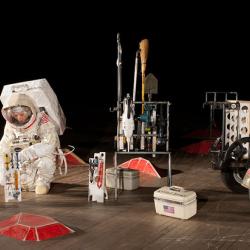 A person dressed in an astronaut suit setting up a display of sculptures that depict the space programs mission to Mars, such as a rover.