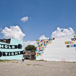 Two buildings beside each other. Both have murals painted on them. One has a light blue background and the text hold tight written on to arms. The other has a white background and magnetic colorful letters.
