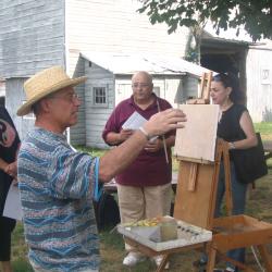 Four people outside of a barn, one of whom has an easel set up and is about to paint