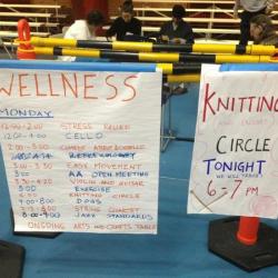 In a school gymnasium, two lists hang from a low pole. One has a schedule of Wellness events and the other advertises a knitting circle.