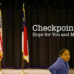 A young person in a blue suit standing in front of a stage with the US flag and the North Carolina flag. Text on the photo reads "Checkpoint: Hope for you and me"