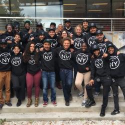 Group of smiling people posing on the steps in black sweaters with "Made in NY" on them
