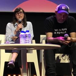 Two people sitting on a stage in front of a table holding microphones. One person is speaking.