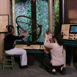 At an exhibit, a young person sits with headphones on and holding onto a brass sculpture. A young child is being assisted with putting on headphones.