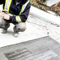 A person in construction gear smiles, leaning over drying concrete with a poem written into it.