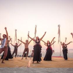 A group of dancers standing in the desert with the sun behind them. They are all in black and are swing pieces of fabrics.