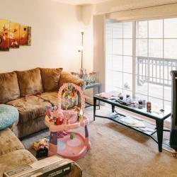A living room with two tan couches and various wall art. There is a sunny sliding door that leads to a balcony. A baby bouncer sits in the middle of the floor.