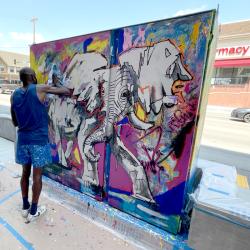 A person stands at a concrete panel painting. The painting is colorful and features the image of an elephant.