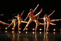 Students in gold outfits dance on a low lit stage