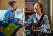Person with a guitar singing along with a child sitting in a hospital bed. Both are in blue.