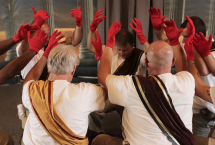 People sitting in a circle in robes with sashes holding up their hands. Their hands have red gloves on them
