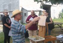 Four people outside of a barn, one of whom has an easel set up and is about to paint