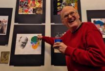 A person in a red sweater smiling and point at a work of art featuring his own face