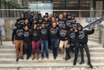 Group of smiling people posing on the steps in black sweaters with "Made in NY" on them