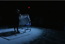 On a darkened stage, a single person stands playing a drum.